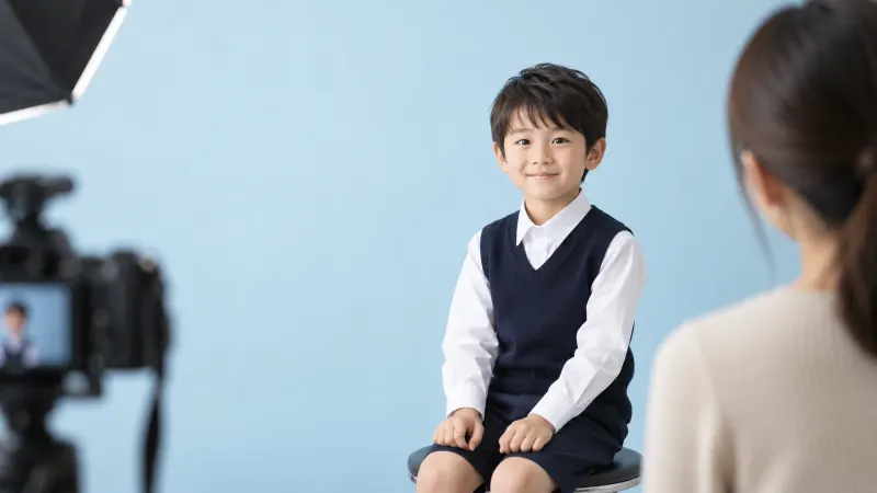 写真館の水色の背景の前で、受験用の白いシャツと紺のベストを着た男の子が証明写真を撮影している様子