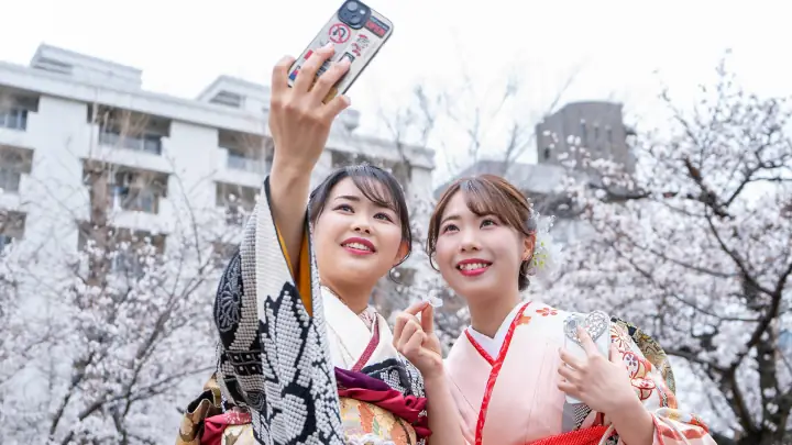 桜の公園で、振袖姿の女性2人がスマートフォンで自撮りする様子